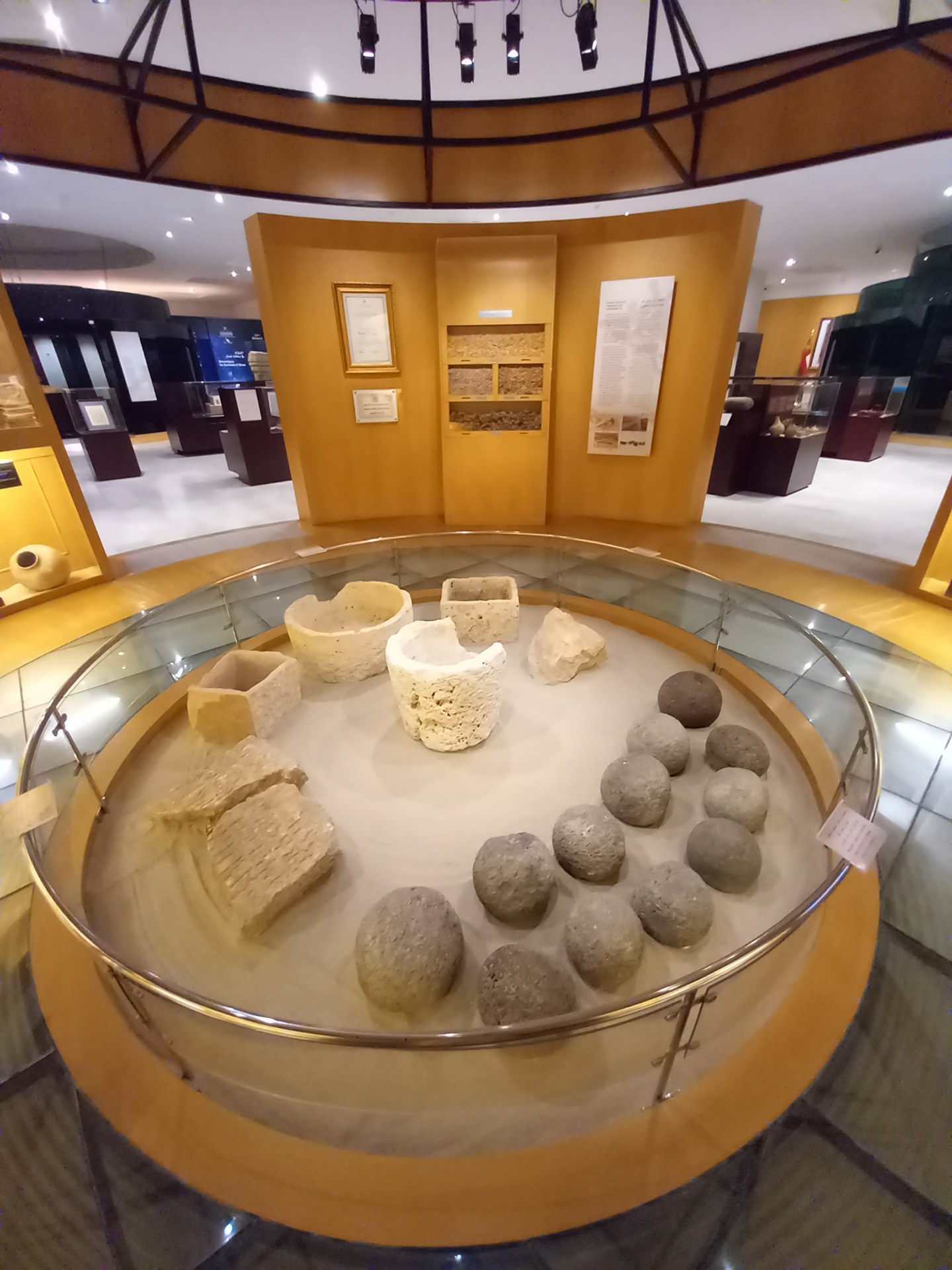 Muzejní expozice muzea země kadidla v salalah s kruhovou vitrínou, kamennými artefakty a žlutými panely, osvětlená stropními reflektory.