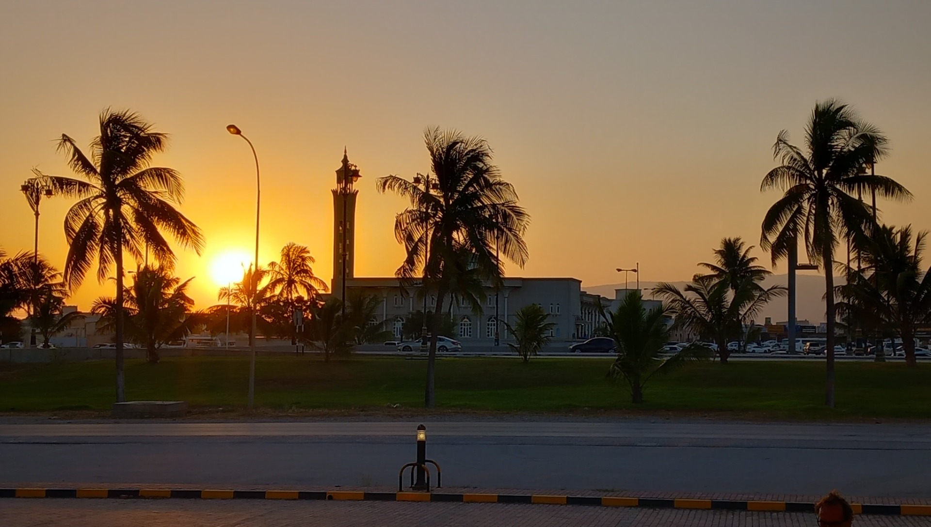 Západ slunce nad Salalah, osvětlená mešita a lampy, siluety palem na pozadí oranžové oblohy.