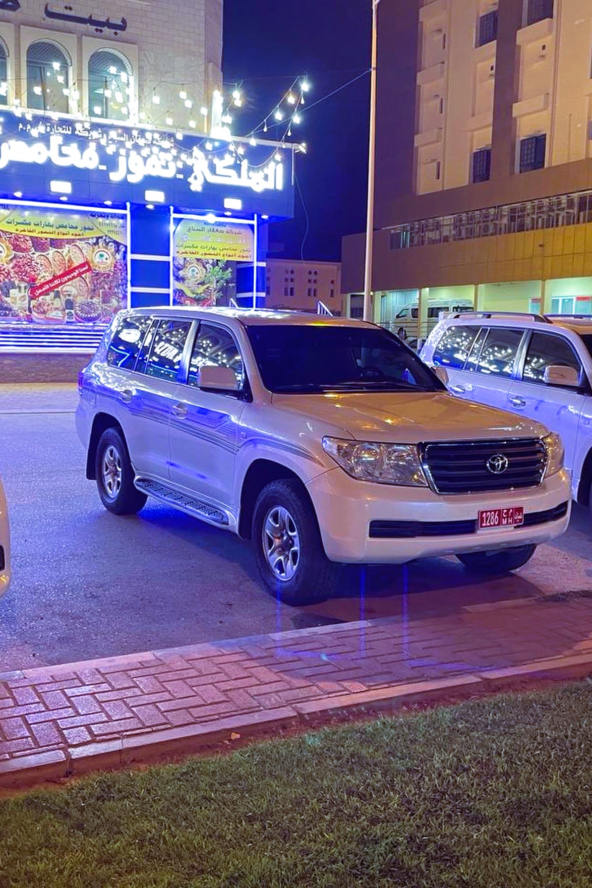 Bílá SUV auta zaparkovaná v noci před osvětlenou budovou v Salalah