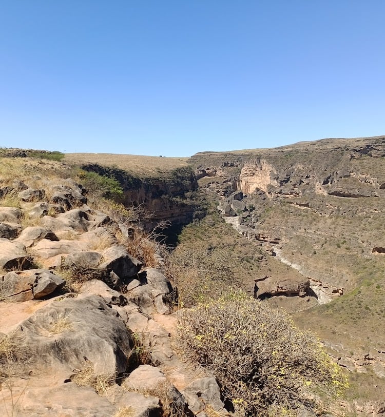 Suchá skalnatá propadlina Taiq cave v oblasti Salalah na jihu Ománu s keři a kamenitými svahy pod jasnou modrou oblohou.