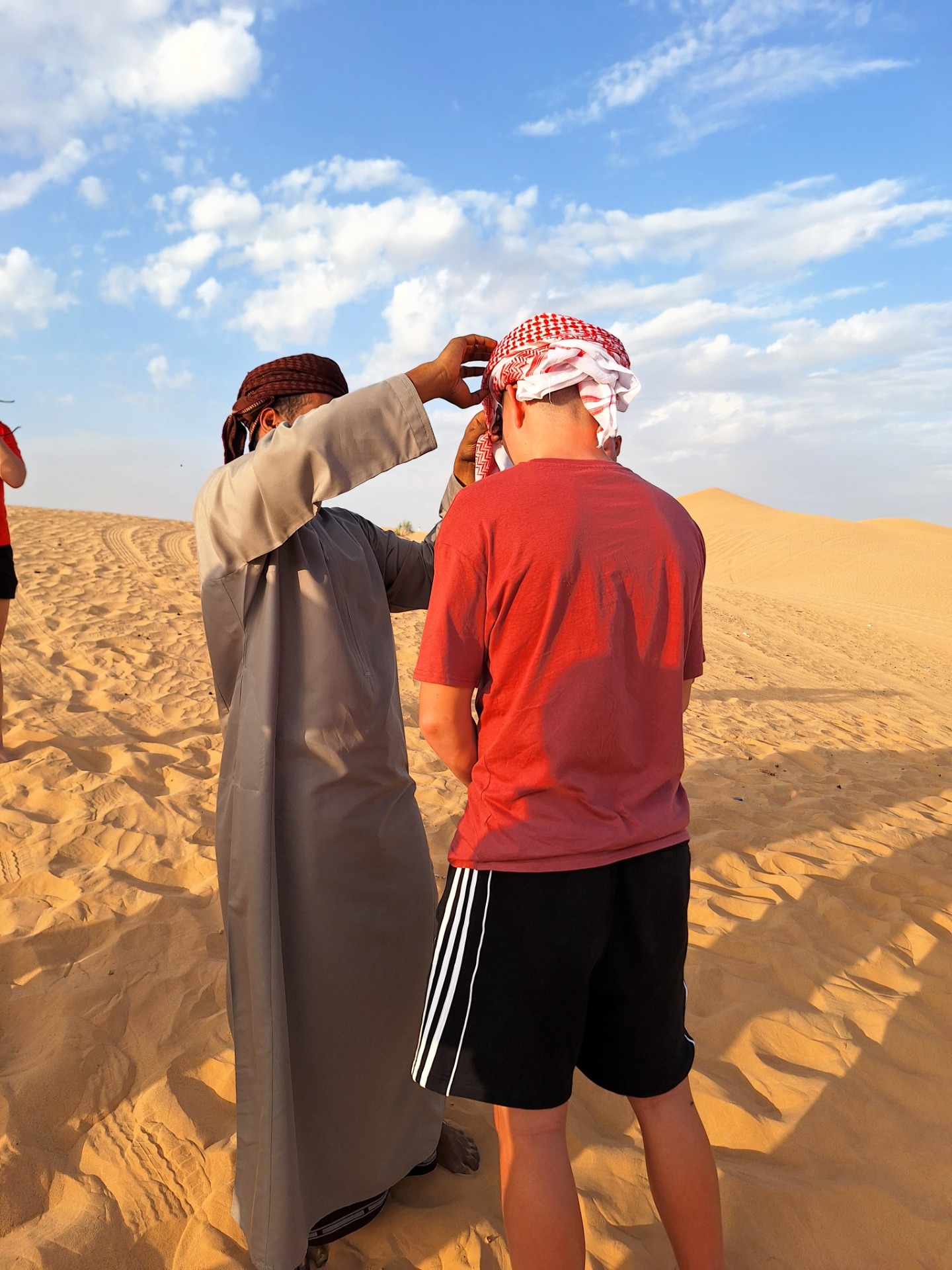 Dva muži v tradičních oděvech si v písečných dunách Rub al khalí, jeden upravuje tradiční mussar šátek proti větru.