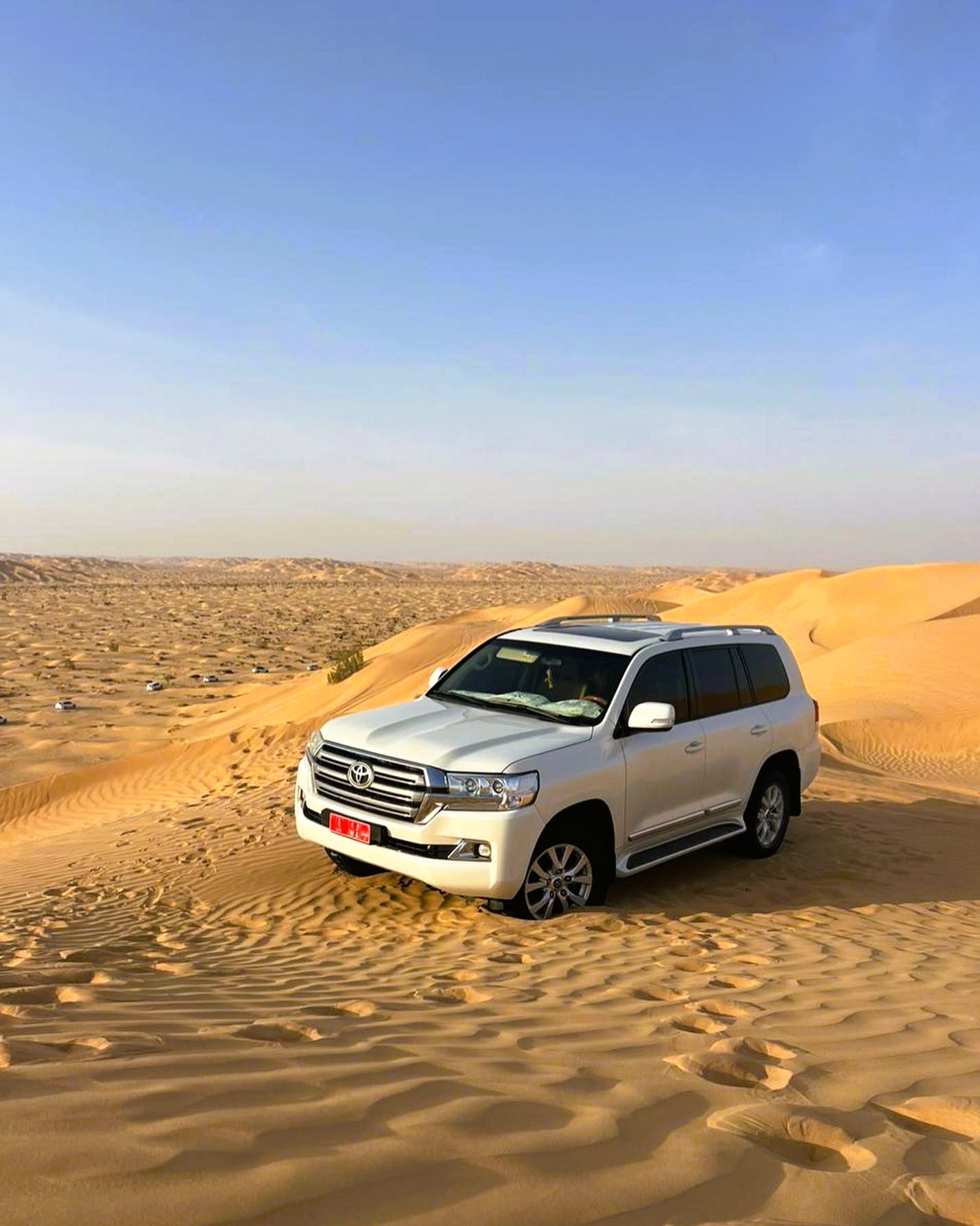 Bílý SUV projíždí písečnými dunami v ománské poušti Rub al Khalí pod jasnou oblohou.