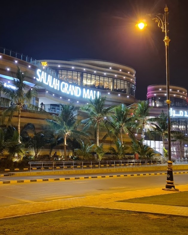 Noční pohled na nákupní centrum Salalah Grand Mall s osvětlenou fasádou, palmy a lampou u silnice.