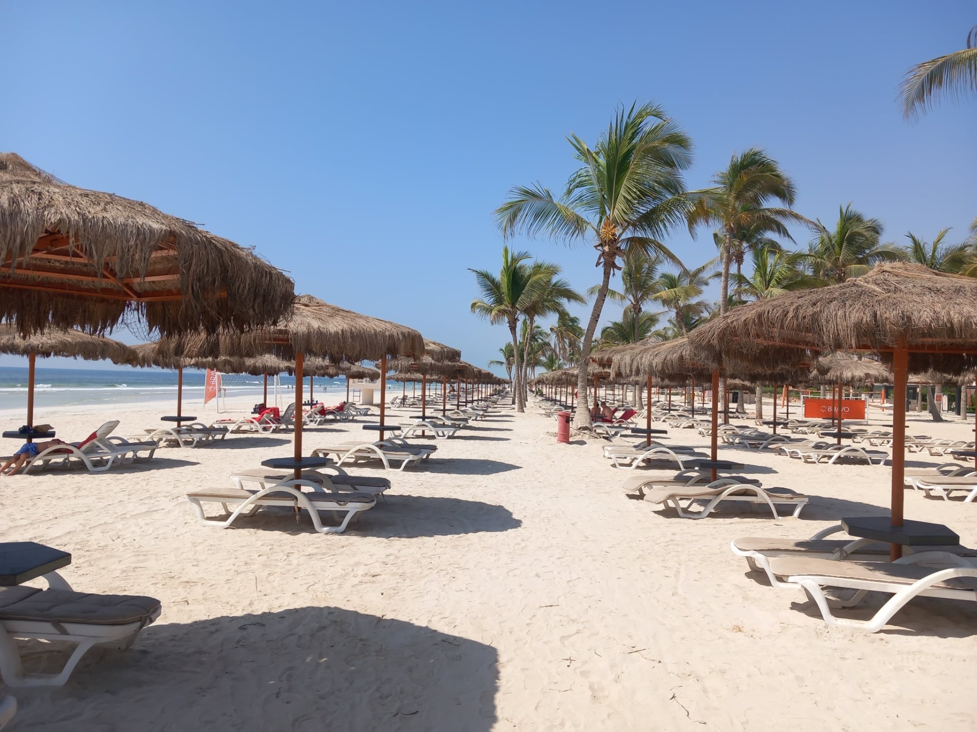 pláž hotelu Rotana salalah s řadou lehátek pod slámovými stínidly, palmy a modré nebe nad mořem.
