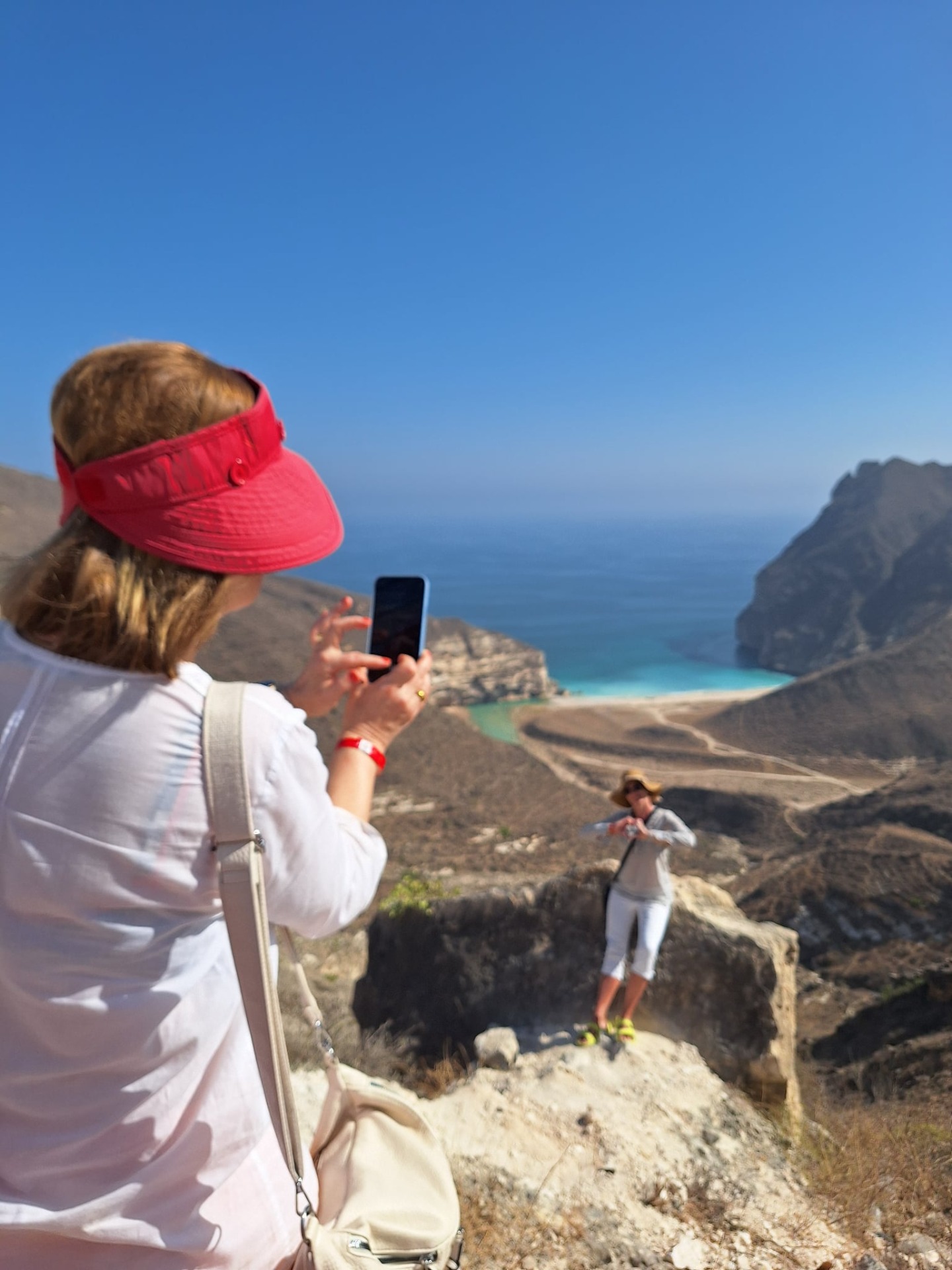 Turistka v červeném kšiltu fotí telefonem vyhlídku na Ztracenou pláž u Salalah a Arabské moře, skalnaté pobřeží a tyrkysovou zátoku.