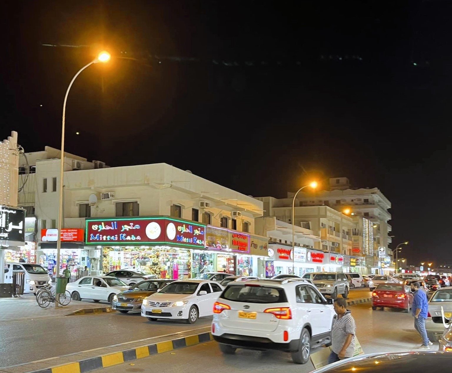 Noční ulice v Salalah s auty, osvětlenými lampami a řadou obchodů s arabskými nápisy.
