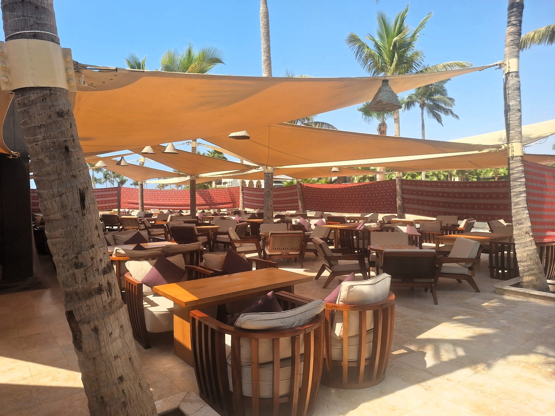 Slunečná terasa na plážovém baru hotelu Salalah Rotana Resort s dřevěnými lehátky, stoly a velkými slunečníky mezi palmami.