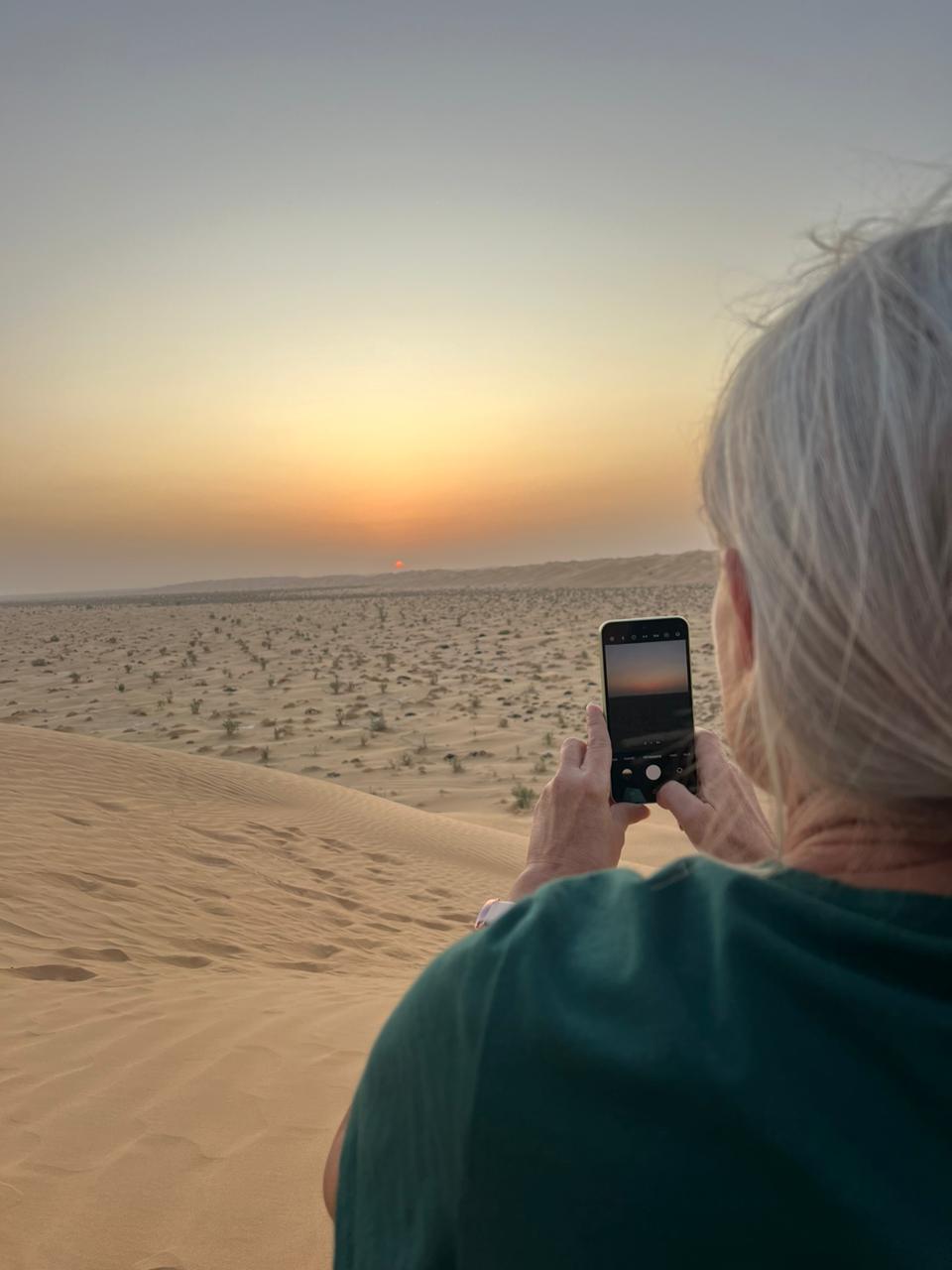 Žena v písečných dunách Rub al khali v Ománu při západu slunce fotí krajinu mobilem, v pozadí poušť a obzor.