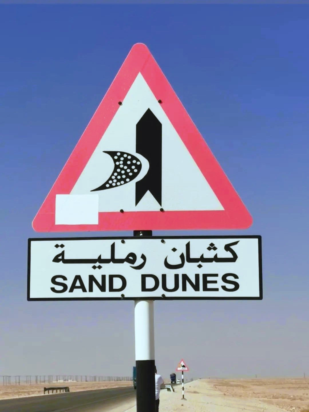 Dopravní značka s nápisem SAND DUNES a arabským textem, červený trojúhelník na pouštní silnici v Ománu